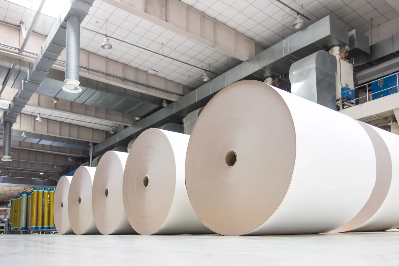 Grandes bobinas de papel industrial almacenadas dentro de una planta de fabricación con maquinaria y área de producción al fondo.
